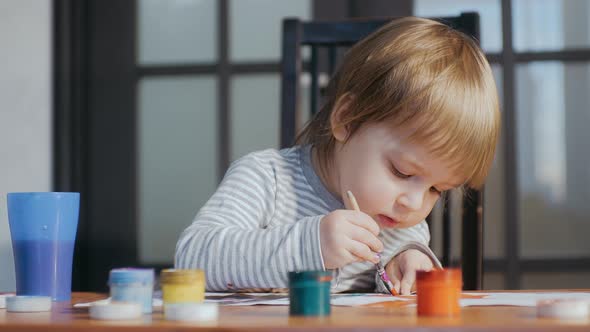 A Small Funny Child of 2-3 Years Old Drawing Using Paints and Brushes Sitting at a Table