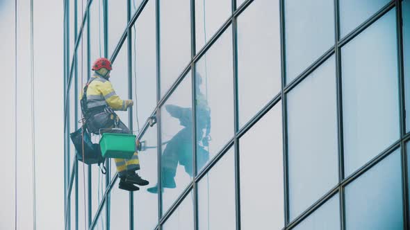 A Man Worker in Yellow Work Clothes Hanging on Ropes and Wipes the Exterior Windows of a Business