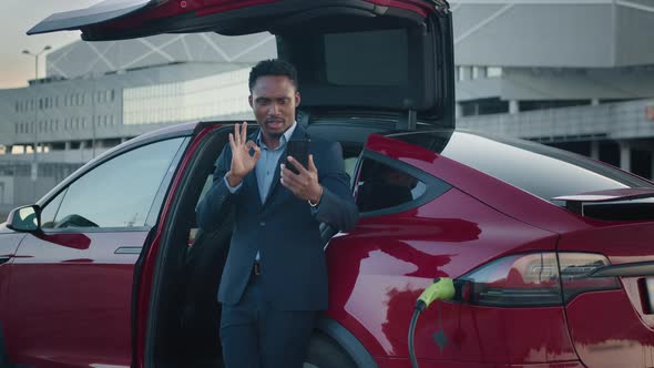 African Man Standing Near Luxury Charging Electric Car and Talking on Mobile