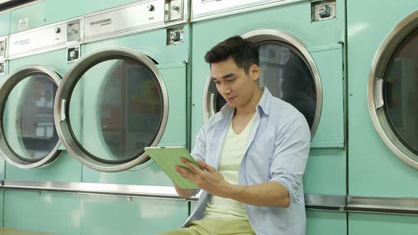 A Young Man sits using his Digital Tablet whilst waiting for his laundry