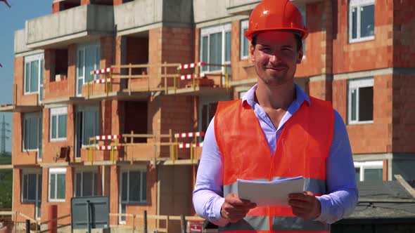 Construction Worker Holds Some Papers and Smiles To Camera in Front of Building Site