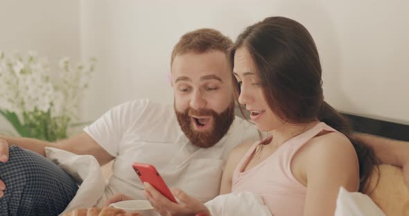 Surprised Man and Woman Looking at Phone Screen and Smiling While Lying on Bed, Cheerful Romantic