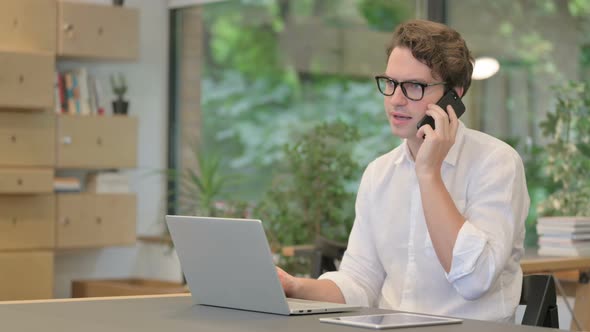 Angry Man Talking on Smartphone While Using Laptop in Modern Office