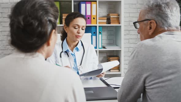 Doctor Woman Consulting Two Elderly Patients Sitting at Table in Office of Clinic Explaining Details