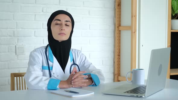 A Doctor in a Hijab and a Medical Uniform Sits at a Table in a Hospital Looking at the Camera with a