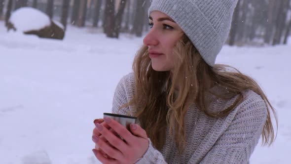 Cute Caucasian Girl in a White Sweater and Hat in a Snowy Forest Park Drinking Hot Tea or Chocolate