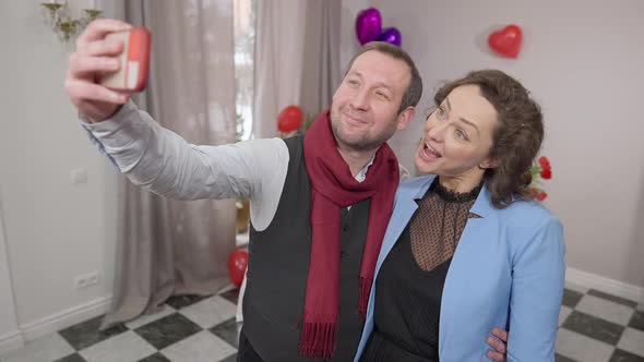 Positive Funny Adult Couple Making Faces Taking Selfie Indoors on Valentine's Day