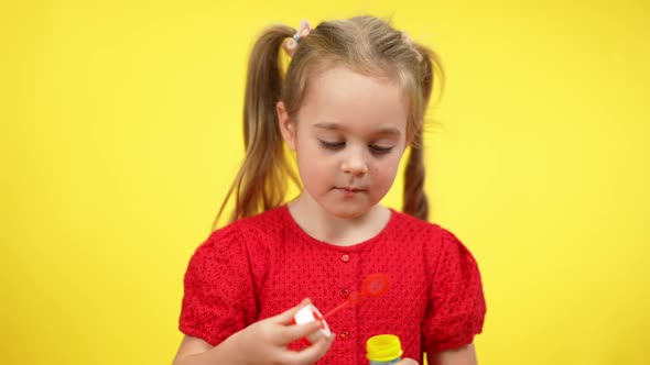 Portrait of Blond Cute Girl with Pigtails Blowing Soap Bubbles at Yellow Background