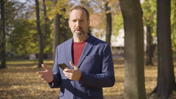 A Middleaged Handsome Caucasian Man Talks to the Camera and Holds a Smartphone in a Park in Fall