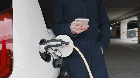 Woman Standing Near Charging an Electric Car and Using Her Mobile Phone