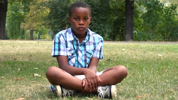 A Young  Black Boy Sits on Grass in a Park and Looks Seriously at the Camera