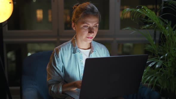 Woman is Sitting in the Armchair and Working on a Laptop at Night or Texting Someone