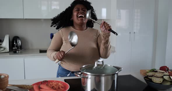Black young african woman having fun cooking and dancing inside kitchen at home