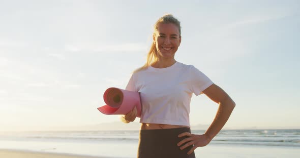 Portrait of caucasian woman holding yoga mat at the beach smiling