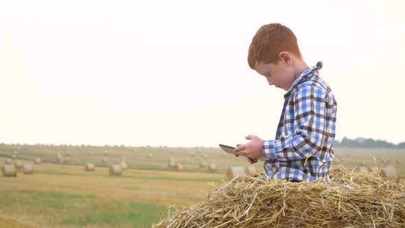A Red-haired Boy on Top of a Straw Bale Holds a Tablet in His Hands on a Background of Hay Bales