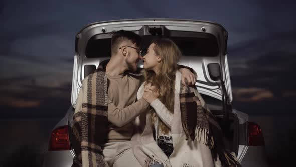 Happy Couple Rubbing Noses Sitting in Car Trunk with Beautiful Summer Twilight at the Background
