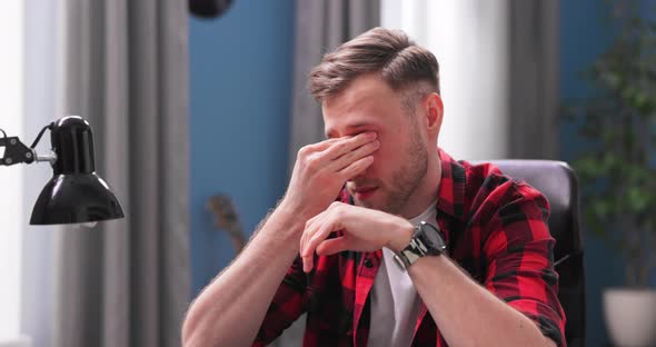 A Handsome Young Man Rubs His Eyes with Fatigue While Working Remotely From Home