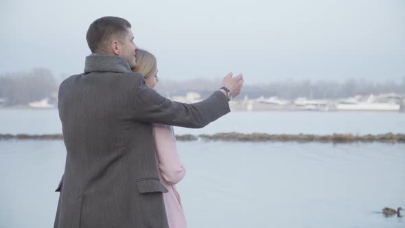 Romantic Caucasian Couple Hugging on Riverbank and Chatting. Back Side View of Younger Man and Older