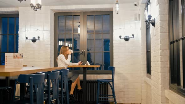 Woman Is Using Phone and Drinking Coffee Alone in Cozy Bar in the Late Evening