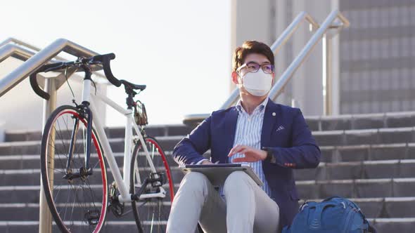 Asian man wearing face mask with laptop sitting on the stairs at corporate park