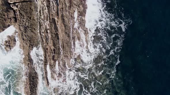 Seawater splashing on rocky coastline with birds flying below, top down view