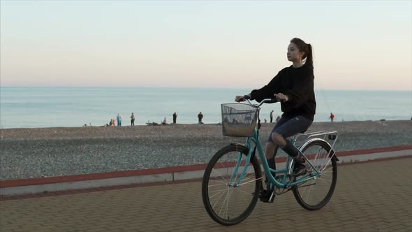 Teenage Girl Is Riding a Bike Along Gravel Beach in Evening Near Sea, Side View