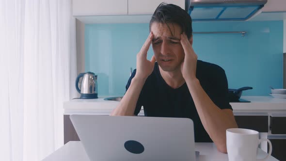 Depressive Young Man Suffers and Has a Headache While Working on a Laptop at Home in the Kitchen