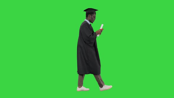 African American Male Student in Graduation Robe Texting on the Phone While Walking with His Diploma