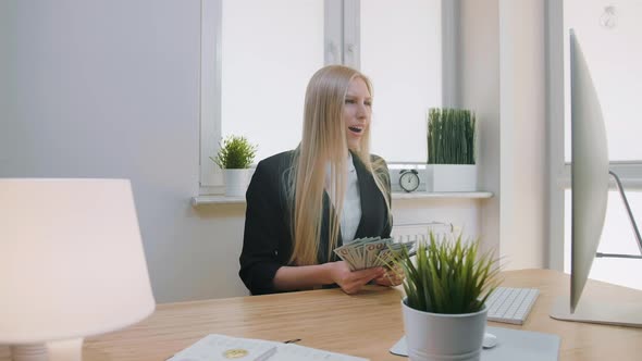 Woman Celebrating Success in Office. Elegant Blond Female Sitting at Workplace Holding in Hands