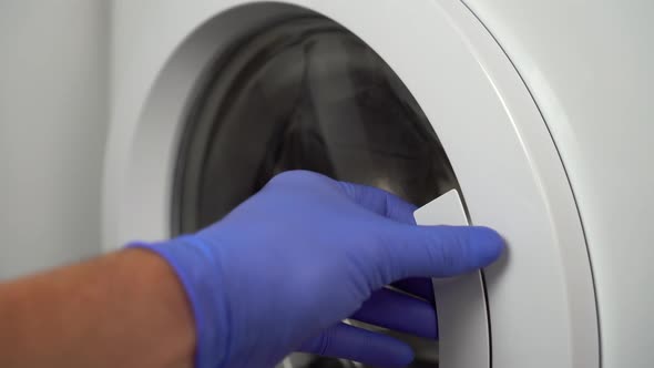 hand in medical blue gloves opens the door of a white washing machine.