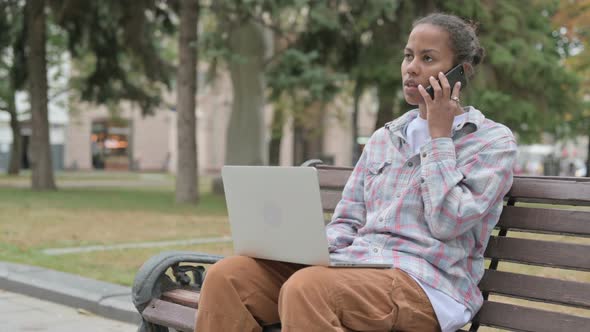 African Woman Talking on Phone and Using Laptop While Sitting Outdoor on Bench