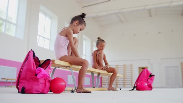 Gymnastics Training  Little Girls Sitting on a Bench in a Sports Hall and Putting on Ballet Shoes