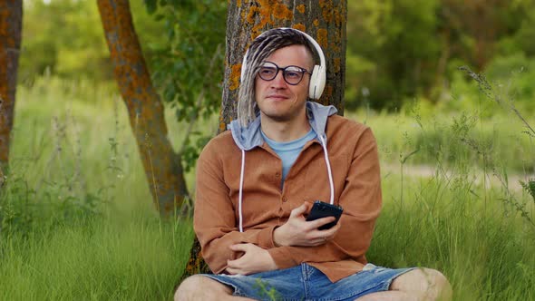 Young Relaxed Man in Glasses Headphones Sitting Near Tree in Green Grass Listening to Music on
