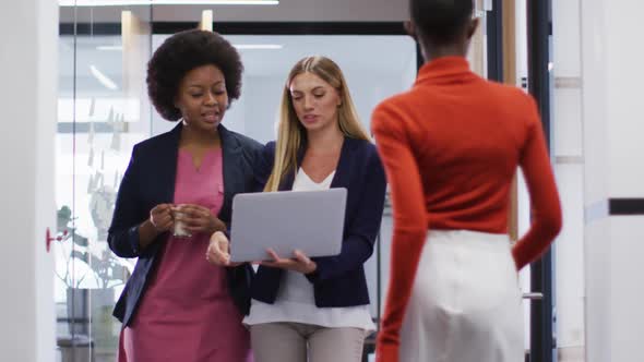 Two diverse female office colleagues with laptop discussing together while walking in office