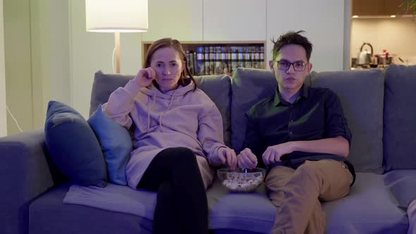 A Young Man in Glasses is Sitting on the Sofa with His Mother Eating Popcorn and Watching TV