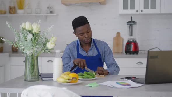 Focused African American Young Man Slicing Cucumber on Cutting Board with Kitchen Knife Sitting at