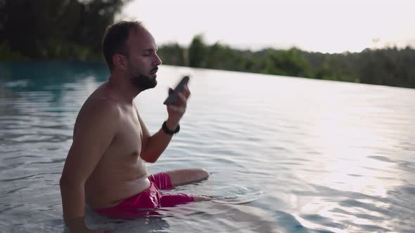 Middle Age Man with a Smartphone Sitting in a Swimming Pool at Sunset and Taking on a Phone