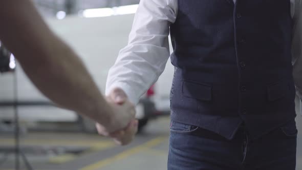 Close-up of Two Caucasian Men Shaking Hands in Car Repair Shop. Male Hands at the Background of