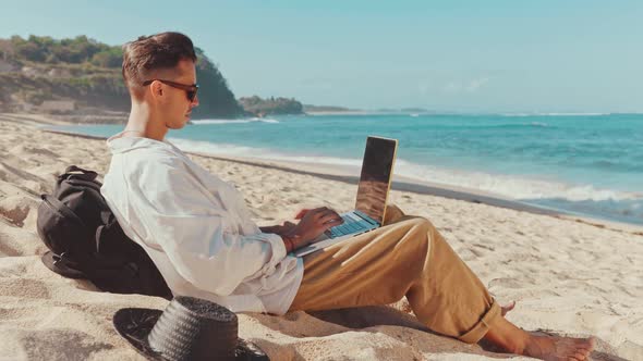 Young Urban Digital Nomad of Millennial Sits on the Beach and Works on Laptop