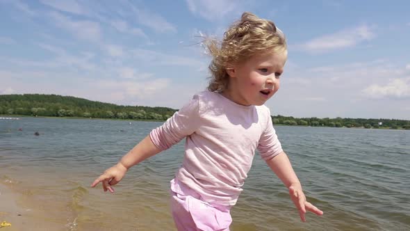 A happy blonde little girl in a pink skirt runs barefoot on a sandy beach.