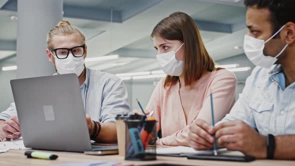 Young Diverse Employees in Protective Masks Working at Notebook Discussing Business Project Sitting