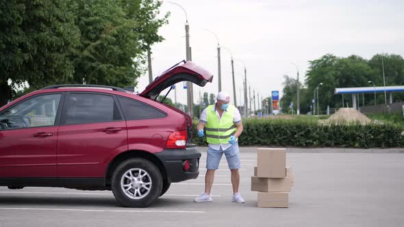 Male Courier, in a Special Vest, Protective Mask, Gloves, Loading Parcels, Putting Cardboard Boxes