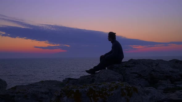 Young Man Sitting, Waits for the Sunrise at the Sea in a Blue Hour. General View