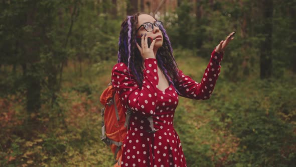 Lost young woman in glasses with backpack talking on mobile phone, standing in summer forest.