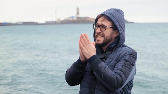 Bearded Man in Glasses Warms Hands Standing Against Blue Sea