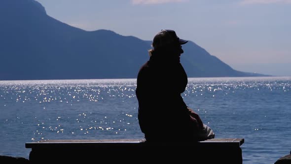 Silhouette of a Lonely Aged Man Sitting on a Bench on Backdrop of a Lake and Mountains