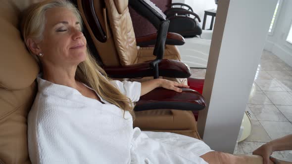 While having her calf massaged, a mature caucasian woman wearing a white robe, sits back and smiles