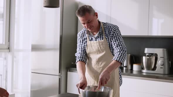 Woman Kissing Her Husband Standing at Table in Kitchen Mixing Ingredients in Bowl