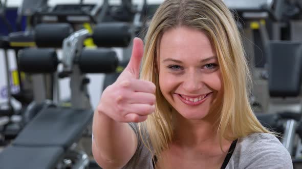 A Young Beautiful Woman Smiles and Shows a Thumb Up To the Camera in a Gym