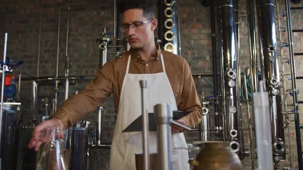 Caucasian man working at distillery checking gin product in flask and holding clipboard